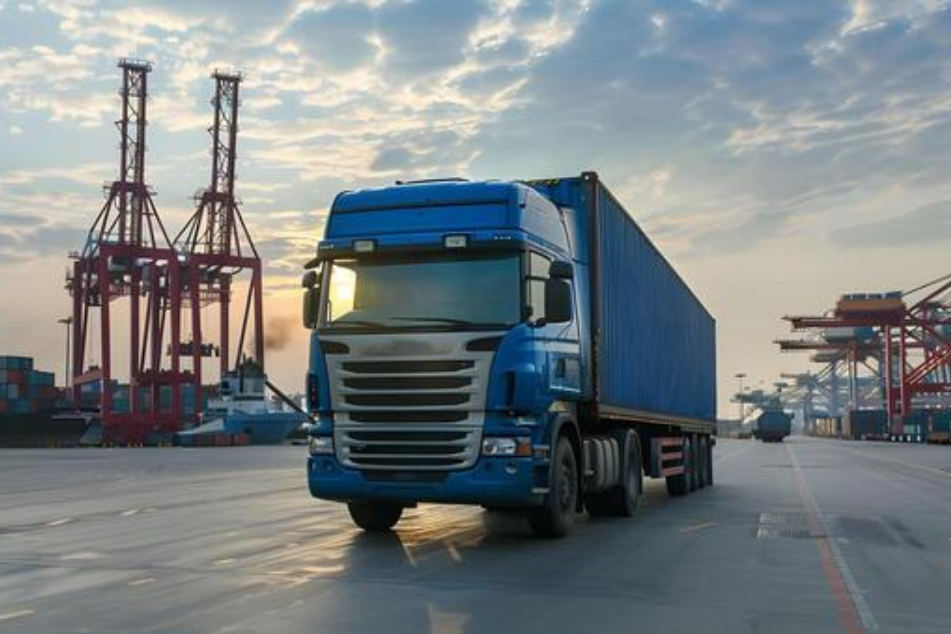 Синий тягач Scania с синим 40-футовым контейнером на асфальте в глубоководном порту, на фоне портовых кранов и рассвета.